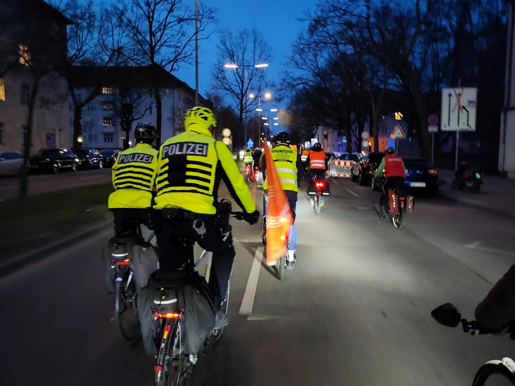 Eine Gruppe von Radfahrenden fahren in der Abenddämmerung eine breite Straße entlang. Im Vordergrund zwei Polizisten, die ebenfalls auf Rädern die Gruppe begleiten. Sie alle tragen leuchtend gelbe Westen.